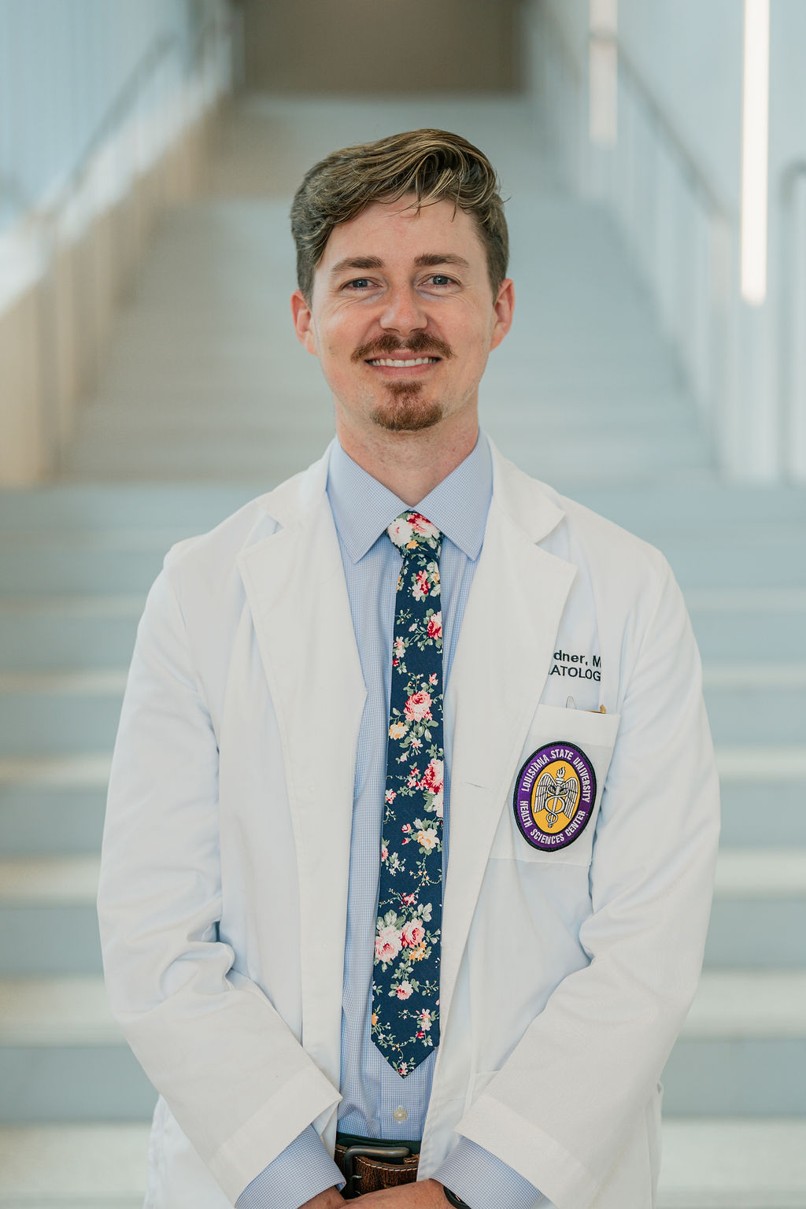 Professional portrait of an individual standing in front of a softly blurred staircase in a bright indoor setting. The individual is wearing a white medical coat over a light blue dress shirt and a dark floral-patterned tie. A Louisiana State University Health Sciences Center patch is visible on the coat pocket, and partial embroidered text including “Dermatology” appears on the chest. The portrait is framed from approximately the knees up, with hands clasped in front. Light-colored stairs and metal railings create a clean, modern background with soft, even lighting and a shallow depth of field.