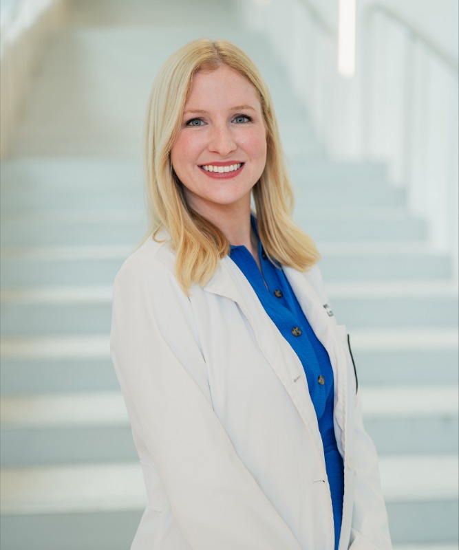 Professional portrait of a person standing in front of a softly blurred staircase in a bright indoor setting. The person is wearing a white lab coat over a blue button-front dress and is turned slightly toward the camera. The image is framed from approximately the knees up, with light-colored stairs and railings visible in the background. The composition is well lit with a shallow depth of field that keeps the background out of focus.