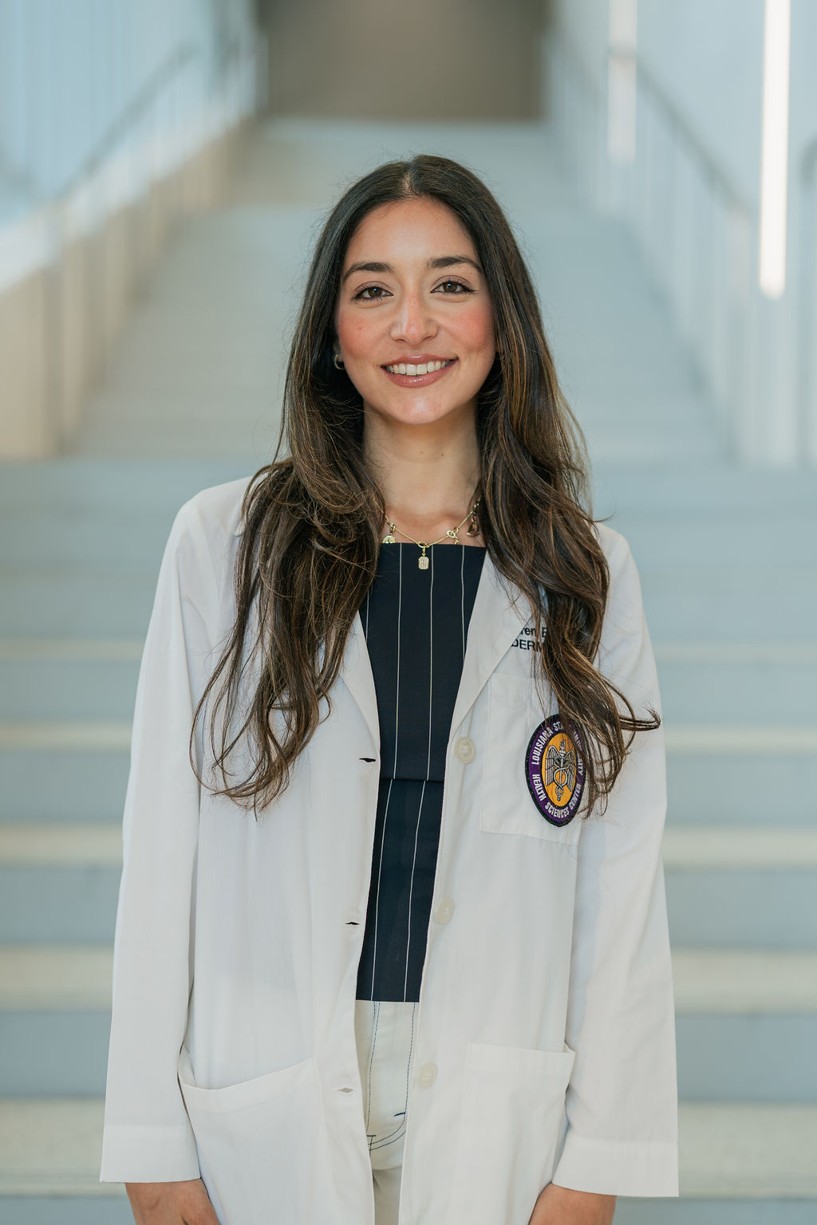 Professional portrait of an individual standing in front of a softly blurred staircase in a bright indoor setting. The individual is wearing a white medical coat over a dark blouse with thin vertical stripes and light-colored trousers. A Louisiana State University Health Sciences Center patch is visible on the coat pocket, and partial embroidered text including “Dermatology” appears on the chest. The portrait is framed from approximately the knees up, with long dark hair and a necklace visible. Light-colored stairs and metal railings create a clean, modern background with soft, even lighting and a shallow depth of field.