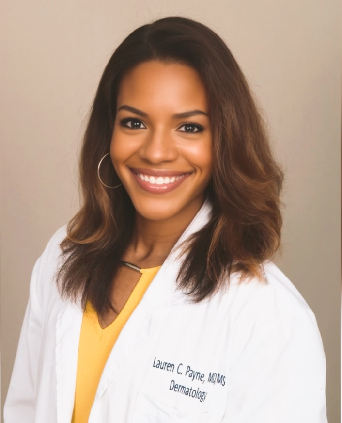 Color studio head‑and‑shoulders portrait of an adult person facing the camera against a warm, neutral background. The person wears a white lab coat over a yellow top, with medium‑length hair styled in loose waves and hoop earrings visible. Lighting is soft and even, creating a polished, professional composition.