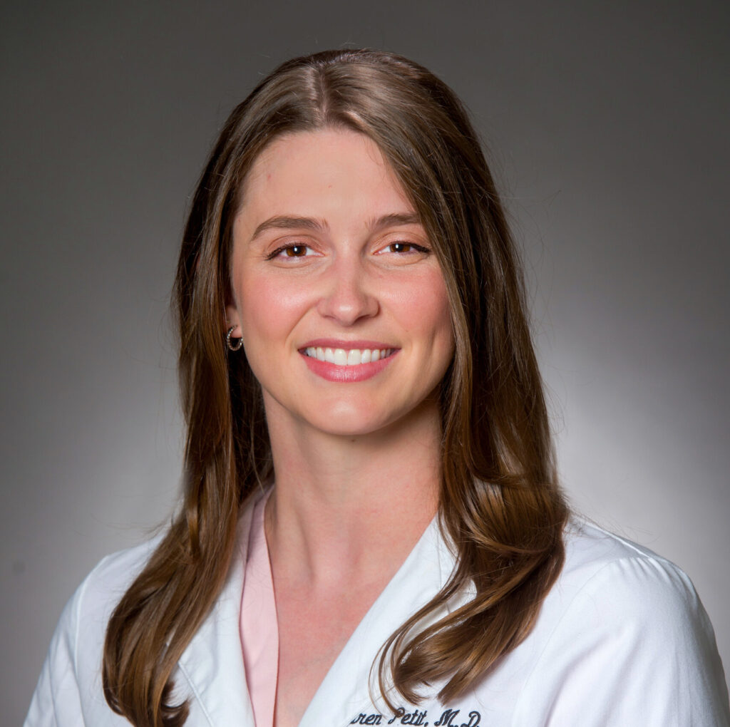  Color studio head-and-shoulders portrait of a person facing the camera against a softly graduated gray background. The person has long, straight hair parted near the center and wears a light-colored coat over a pale top. Lighting is even and professional, with clear facial details and a formal, clean composition.