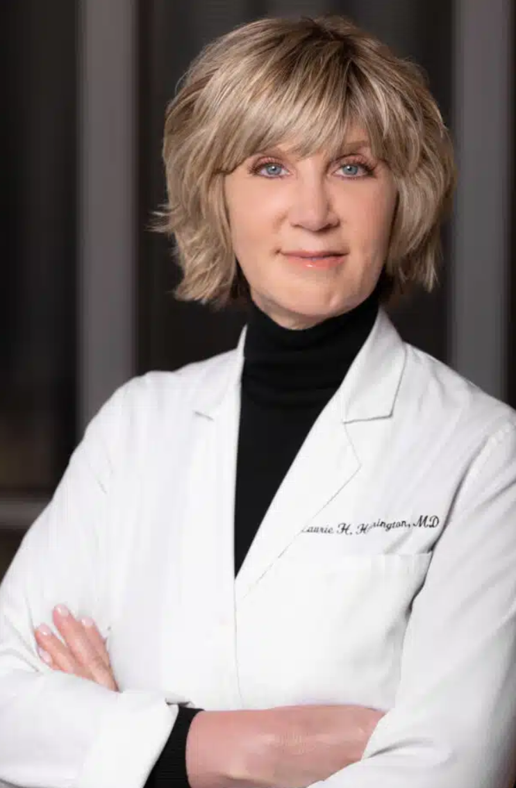 Color studio portrait of an adult person shown from the chest up, facing the camera. The person wears a white lab coat over a dark high‑neck top, with arms crossed. Hair is short and layered, and the background is softly blurred with vertical dark and light tones, creating a professional, clinical setting.