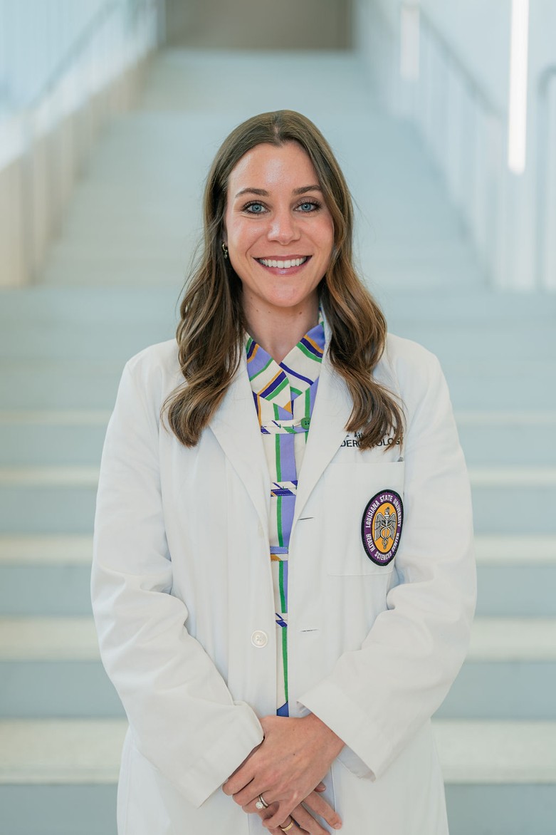 Professional portrait of an individual standing in front of a softly blurred staircase in a bright indoor setting. The individual is wearing a white medical coat over a patterned blouse with geometric shapes in blue, green, purple, and gold tones. A Louisiana State University Health Sciences Center patch is visible on the coat pocket, and partial embroidered text reading “Dermatology” appears on the chest. The portrait is framed from approximately the waist up, with hands clasped in front and light-colored stairs and railings creating a clean, modern background.