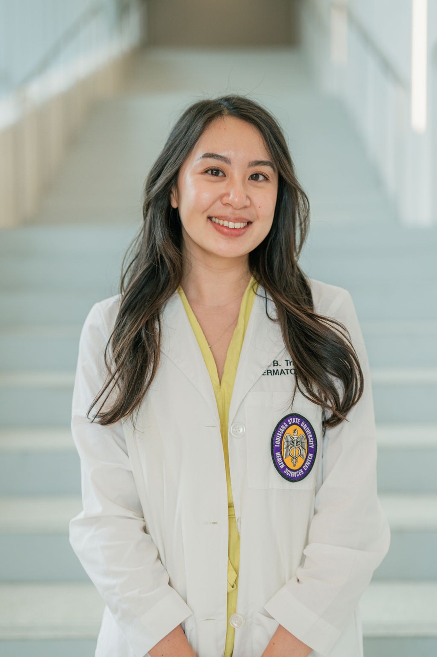 Professional portrait of an individual standing in front of a softly blurred staircase in a bright indoor setting. The individual is wearing a white medical coat over a light yellow dress or blouse. A Louisiana State University Health Sciences Center patch is visible on the coat pocket, and partial embroidered text including “Dermatology” appears on the chest. The portrait is framed from approximately the knees up, with hands resting together in front. Light-colored stairs and metal railings create a clean, modern background with soft, even lighting and a shallow depth of field.