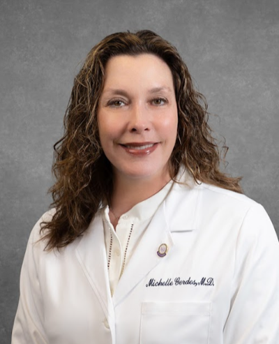 Color studio portrait of an adult person shown from the waist up, facing the camera against a textured gray background. The person wears a white lab coat over a light blouse; the lab coat has small embroidered text on the chest and a pin on the lapel. Hair is medium length and wavy. Lighting is even and professional, creating a clean, formal composition.