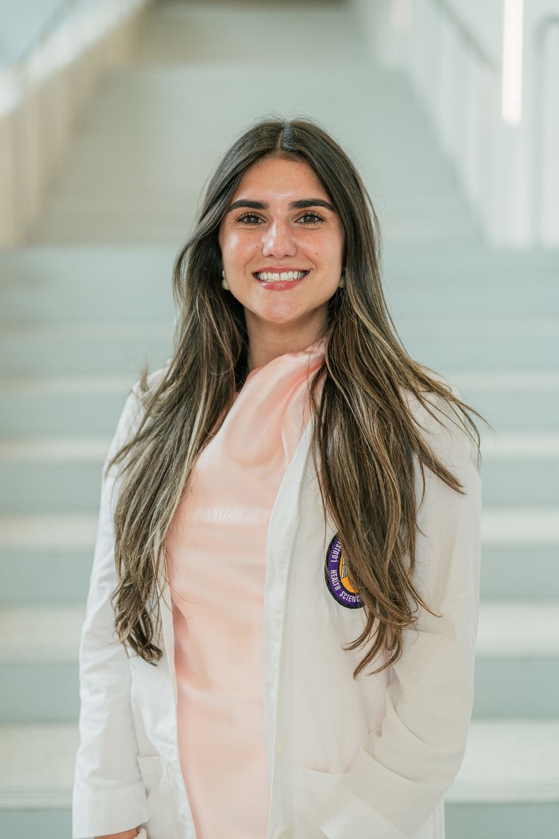 Professional portrait of an individual standing in front of a softly blurred staircase in a bright indoor setting. The individual is wearing a white medical coat over a light pink dress or blouse. Long dark hair falls past the shoulders. A partially visible Louisiana State University Health Sciences Center patch appears on the coat pocket. The portrait is framed from approximately the knees up, with the body angled slightly toward the camera. Light-colored stairs and metal railings create a clean, modern background with soft, even lighting and a shallow depth of field.