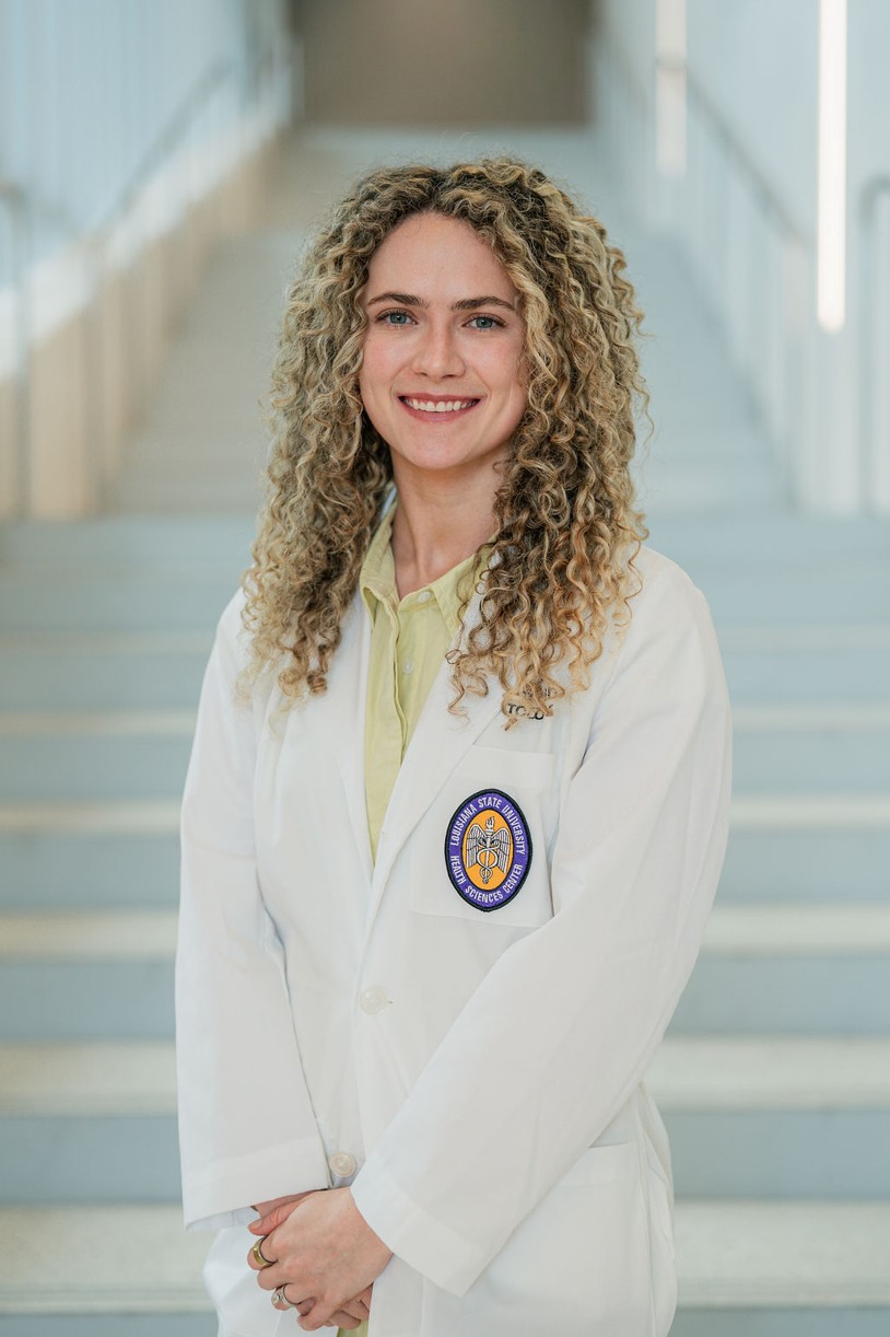 Professional portrait of an individual standing in front of a softly blurred staircase in a bright indoor setting. The individual is wearing a white medical coat over a light green collared dress or blouse. A Louisiana State University Health Sciences Center patch is visible on the coat pocket. The portrait is framed from approximately the knees up, with hands clasped in front. Light-colored stairs and metal railings create a clean, modern background with soft, even lighting and a shallow depth of field.