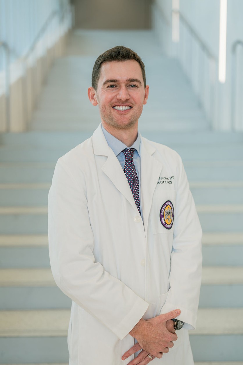 Professional portrait of an individual standing in front of a softly blurred staircase in a bright indoor setting. The individual is wearing a white medical coat over a light blue dress shirt and a patterned tie. A Louisiana State University Health Sciences Center patch is visible on the coat pocket, and partial embroidered text including “Dermatology” appears on the chest. The portrait is framed from approximately the knees up, with hands clasped in front. Light-colored stairs and railings create a clean, modern background with soft, even lighting and a shallow depth of field.