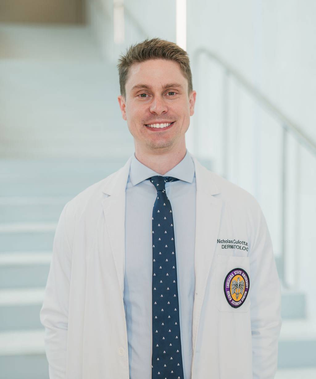 Professional portrait of a person standing in front of a softly blurred staircase in a bright indoor setting. The person is wearing a white medical coat over a light blue dress shirt with a patterned blue tie and blue trousers. Visible text on the coat includes “Dermatology,” along with a Louisiana State University Health Sciences Center patch on the chest pocket. The image is framed from approximately the knees up, with the person facing the camera and the staircase and handrails softly out of focus in the background.
