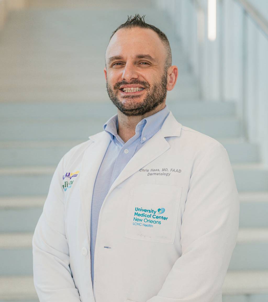 Professional portrait of a person standing in front of a softly blurred staircase in a bright indoor setting. The person is wearing a white medical coat over a light blue collared shirt and is positioned at a slight angle to the camera with hands clasped in front. Visible text on the coat includes “University Medical Center New Orleans” and medical credentials embroidered above the chest pocket. The image is framed from approximately the knees up, with light-colored stairs and railings forming the background.