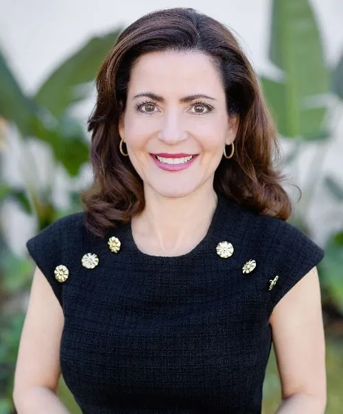 Head-and-shoulders portrait of a person with shoulder-length hair wearing a sleeveless black textured top accented with gold decorative buttons near the neckline. The person faces the camera, with large green leaves softly blurred in the background.