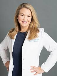Color studio portrait of an adult person shown from the waist up, standing with hands on hips against a neutral gray background. The person wears a white lab coat over a dark top. Hair is shoulder length and styled neatly. Lighting is even and professional, creating a confident, polished composition.