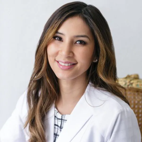Color studio head‑and‑shoulders portrait of an adult person facing the camera against a light, neutral background. The person has long, wavy hair worn down and wears a white lab coat over a patterned top. Lighting is soft and even, with a clean, professional composition and a wicker chair partially visible in the background.