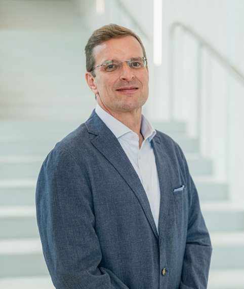 Professional portrait of a person standing in front of a softly blurred staircase in a bright indoor setting. The person is wearing a blue blazer over a light-colored button-down shirt and eyeglasses. The portrait is framed from approximately the knees up, with the person positioned at a slight angle to the camera and hands clasped in front. Light-colored stairs and railings create a clean, modern background with a shallow depth of field.