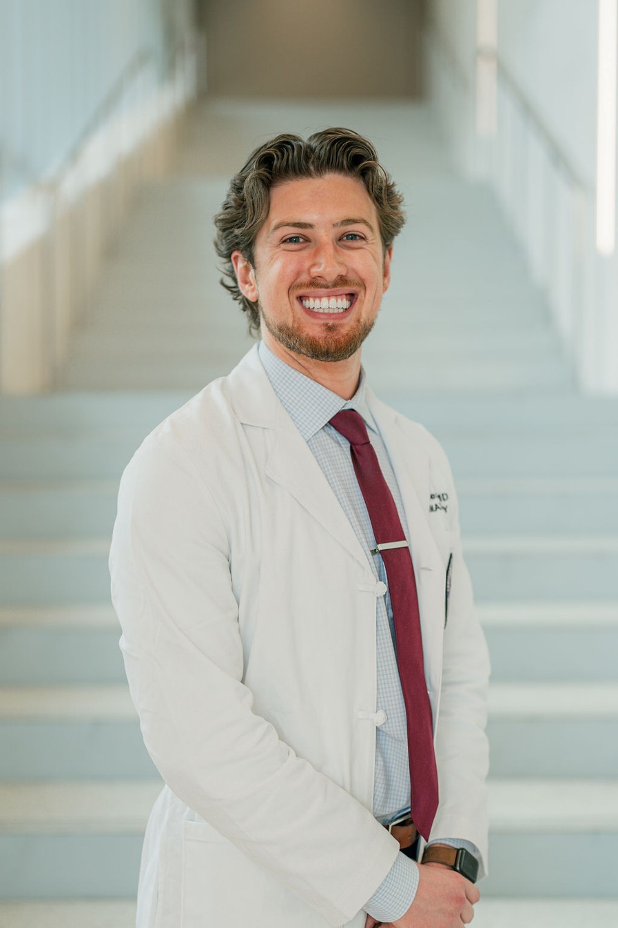 Professional portrait of an individual standing in front of a softly blurred staircase in a bright indoor setting. The individual is wearing a white medical coat over a light-colored checkered dress shirt and a burgundy tie with a tie clip. The portrait is framed from approximately the knees up, with hands clasped in front. Partial embroidered text is visible on the coat, and a watch is worn on one wrist. Light-colored stairs and metal railings create a clean, modern background with soft, even lighting and a shallow depth of field.