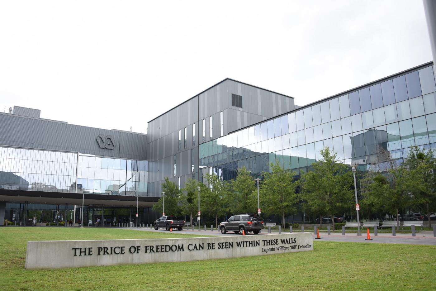 Exterior of a modern VA medical center with reflective glass buildings and a stone monument in the foreground on a grassy lawn.