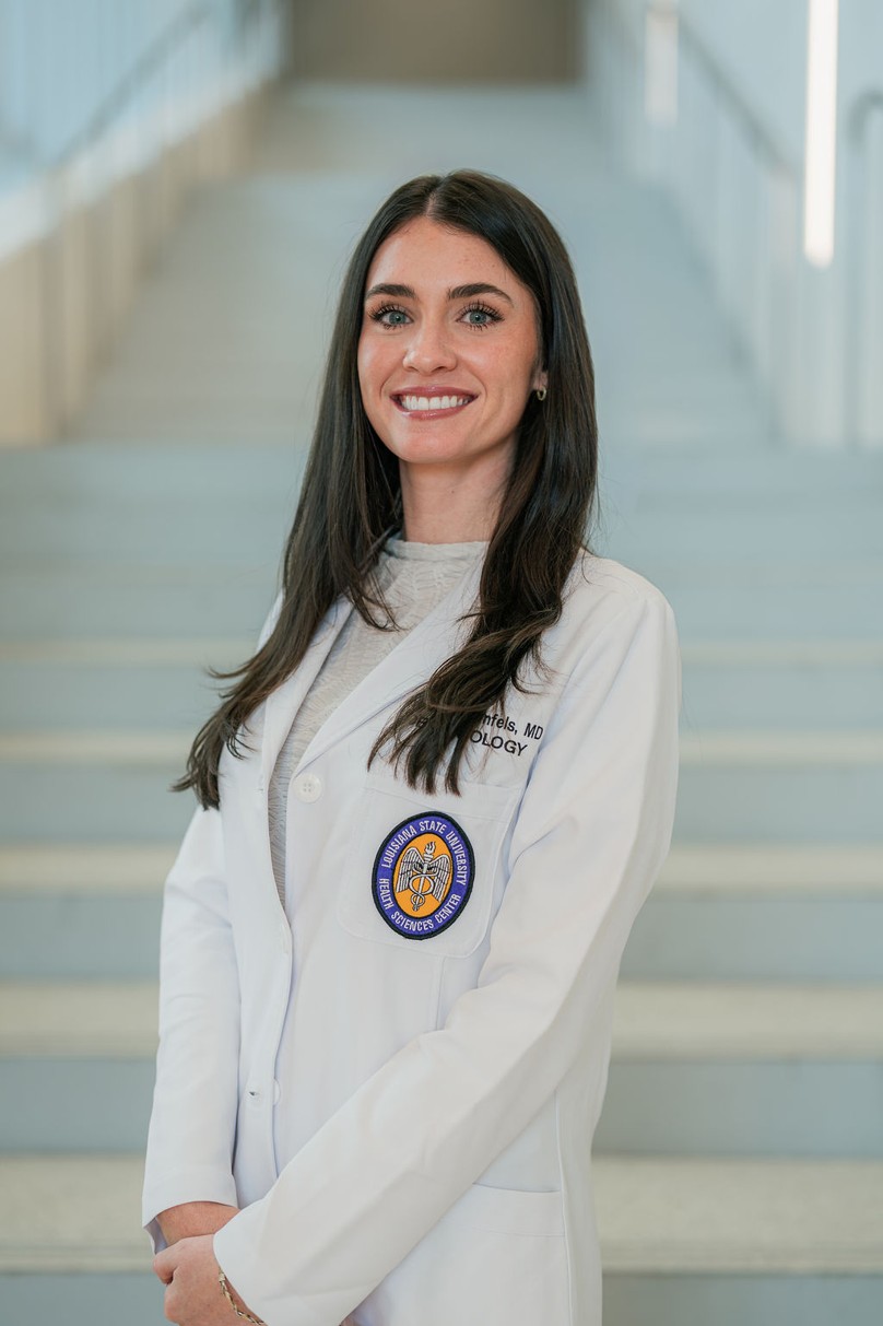 Professional portrait of an individual standing in front of a softly blurred staircase in a bright indoor setting. The individual is wearing a white medical coat over a light-colored top. A Louisiana State University Health Sciences Center patch is visible on the coat pocket, and partial embroidered text including “MD” and “Dermatology” appears on the chest. The portrait is framed from approximately the knees up, with long dark hair and hands clasped in front. Light-colored stairs and metal railings create a clean, modern background with soft, even lighting and a shallow depth of field.