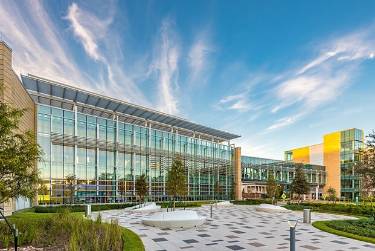 Modern glass hospital building with landscaped courtyard, walkways, and benches under a blue sky.