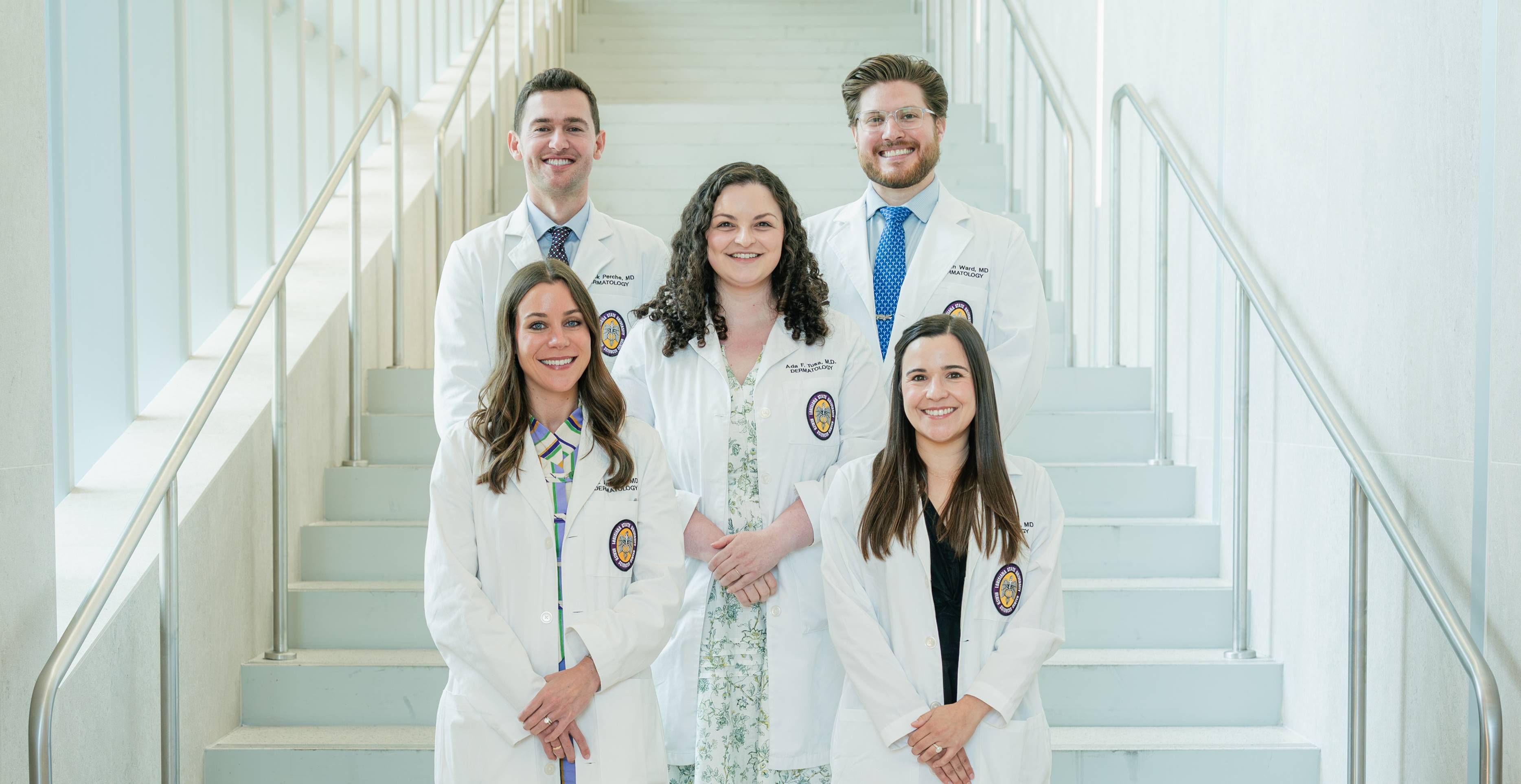 Group portrait of five individuals wearing white medical coats standing on a wide staircase inside a bright, modern building. The group is arranged in three rows, with two people standing in front, one in the middle, and two at the back. Some individuals are wearing professional attire, including dresses, a patterned outfit, dress shirts, and ties beneath their coats. Circular institutional patches are visible on the coats. Metal handrails line both sides of the staircase, and the light-colored walls and steps create a clean, symmetrical background. The image is evenly lit and composed in a formal, professional style.