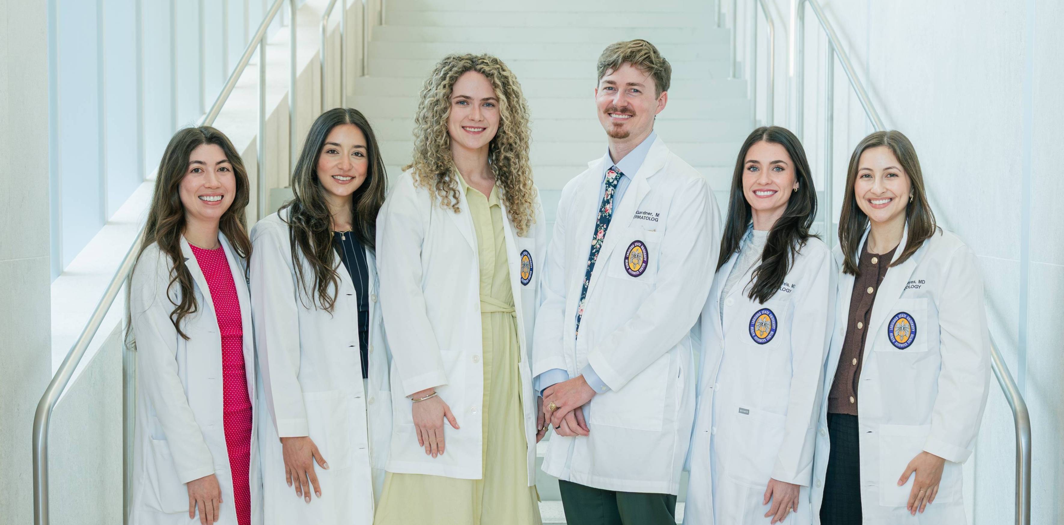 Group portrait of six individuals wearing white medical coats standing together on a wide staircase inside a bright, modern building. The group is arranged in a single row across the lower steps, with various professional outfits visible beneath the coats, including dresses, trousers, dress shirts, and a patterned tie. Circular institutional patches are visible on several coat pockets. Metal handrails frame both sides of the staircase, and the light-colored walls and steps create a clean, symmetrical background. The image is evenly lit and composed in a formal, professional style.