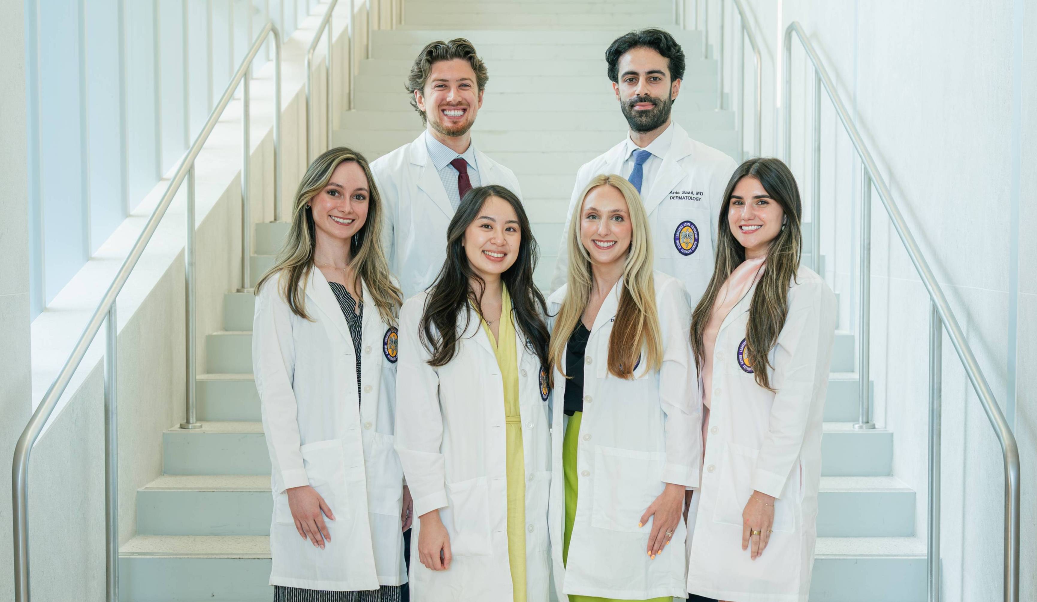 Group portrait of six individuals wearing white medical coats standing on a staircase inside a bright, modern building. The group is arranged in two rows, with four individuals on the lower steps and two on the steps behind them. Professional attire is visible beneath the coats, including dresses, skirts, dress shirts, ties, and trousers. Several coats display Louisiana State University Health Sciences Center patches on the chest pockets. Metal handrails line both sides of the staircase, and the light-colored walls and steps create a clean, symmetrical background. The image is evenly lit and professionally composed.