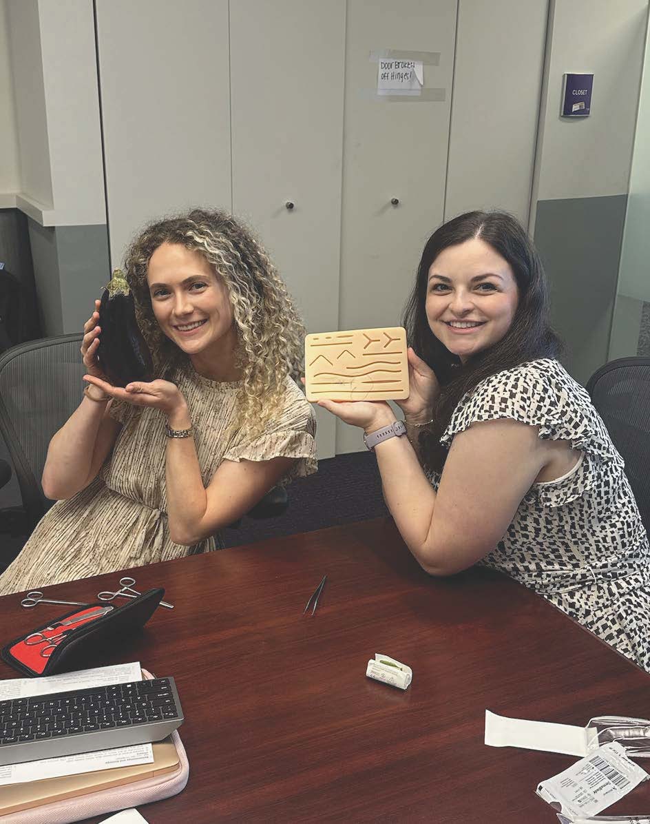 “Two smiling women sit side by side at a conference table in an office, posing with handmade items. The woman on the left holds a dark, textured object in her hands, while the woman on the right holds a small rectangular wooden piece engraved with symbols. Papers and office supplies rest on the table, with cabinets behind them.”