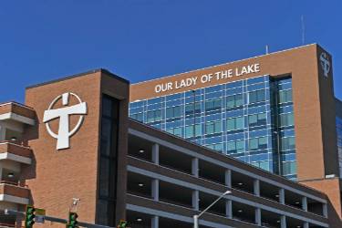 Exterior of Our Lady of the Lake hospital, a multi‑story brick and glass building with a large cross symbol and the hospital name displayed at the top.