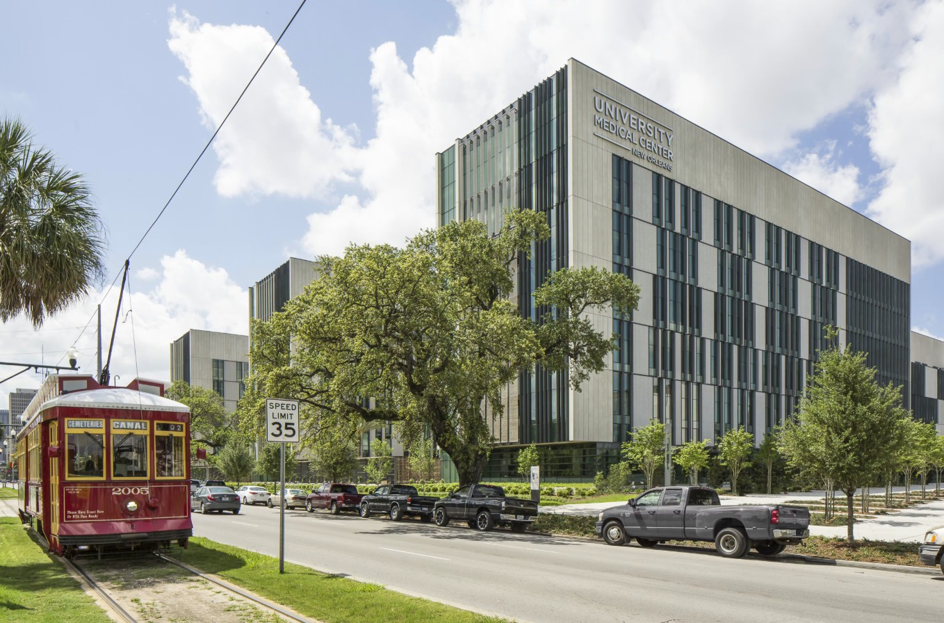 “University Medical Center building along a city street, with a red streetcar in the foreground and cars parked along the road on a sunny day.”