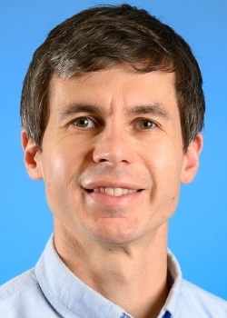 Professional head-and-shoulders portrait of a person with short dark hair wearing a light blue collared shirt, photographed against a solid blue background. The person is facing the camera, and the image is tightly cropped around the face and upper shoulders.