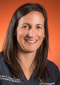 Professional head-and-shoulders portrait of a person with long dark hair wearing dark blue medical scrubs, photographed against a solid orange background. The person is facing the camera, and the image is closely cropped around the face and upper torso. Small white text identifying a medical affiliation is visible on the scrub top near the shoulders and chest.