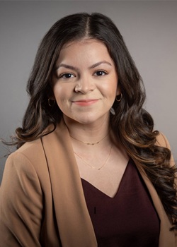 Professional head-and-shoulders portrait of a person with long, dark wavy hair, wearing a tan blazer over a dark burgundy top. The person is facing the camera against a plain gray studio background. The image is closely cropped around the head and upper torso, with soft, even lighting and small hoop earrings and a delicate necklace visible.