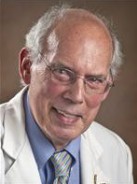 Head-and-shoulders professional portrait of a person wearing a white coat, light blue shirt, and patterned tie. The person has glasses and gray hair around the sides of a balding head. The image is cropped closely to the face and upper shoulders, with a soft brown studio-style background.