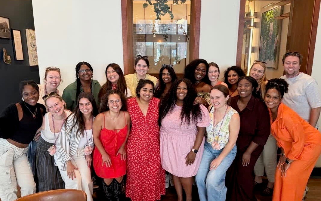 Group photo of approximately 18 people gathered indoors at a restaurant or event venue. The group is arranged in two rows, with individuals standing and crouching together for a posed photo. Participants wear a variety of colorful outfits, including dresses, casual tops, and a bright orange outfit. The setting features wood-framed windows, artwork on the walls, and warm indoor lighting.