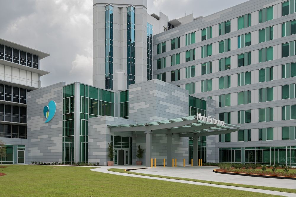 Exterior view of a modern multi-story medical facility with gray and white paneling, large green-tinted windows, and a covered main entrance. A glass canopy extends over the circular drop-off area, with the words “Main Entrance” displayed above the entrance. A landscaped lawn and walkway are visible in the foreground under an overcast sky.