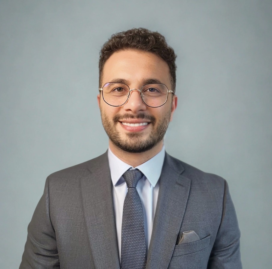 A person wearing a gray suit jacket, white dress shirt, and textured dark tie stands against a plain light-blue backdrop. A pocket square is neatly placed in the jacket’s breast pocket, and the individual has short, curly hair.