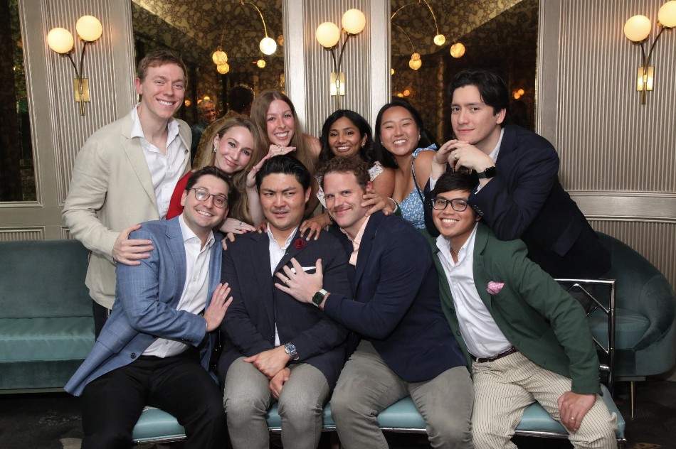 A group of people pose together in a stylish lounge with teal seating, paneled walls, and warm globe lighting. Several individuals sit on a bench at the front while others stand closely behind them, with many placing hands on friends’ shoulders in a playful, celebratory manner. The setting appears festive and social, with everyone dressed in semi-formal attire.